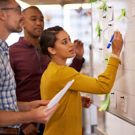 woman in a mustard yellow cardigan writing on a white board with people standing next to her